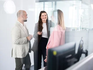 Kollegiale Abstimmung im Arbeitsbereich Drei Personen in Business-Kleidung stehen in einem Büro und unterhalten sich. Im Vordergrund ist ein Computermonitor zu sehen.