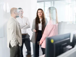 Teamgespräch im Bürobereich Vier Personen in Business-Kleidung stehen in einem Büro und unterhalten sich. Zwei Männer stehen links, zwei Frauen rechts, eine davon hält ein Smartphone. Im Vordergrund rechts ist ein Computermonitor sichtbar.