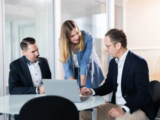 Analyse und Austausch am Laptop Drei Personen in Business-Kleidung sind in einem Büro um einen Tisch versammelt. Eine Person steht und zeigt auf den Laptop-Bildschirm, während die anderen beiden sitzen und aufmerksam zuschauen.