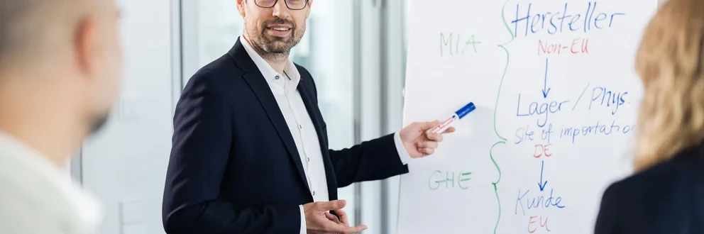 Präsentation im Büro Ein Mann im Anzug hält eine Präsentation für zwei weitere Personen und nutzt dabei ein Flipchart mit Stichpunkten zu Import- und Lagerprozessen.
