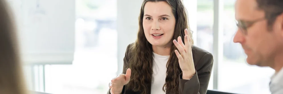 Strategiemeeting mit Fokus auf Erfolg Drei Personen im Gespräch an einem Tisch mit Laptop und Whiteboard im modernen Büro.